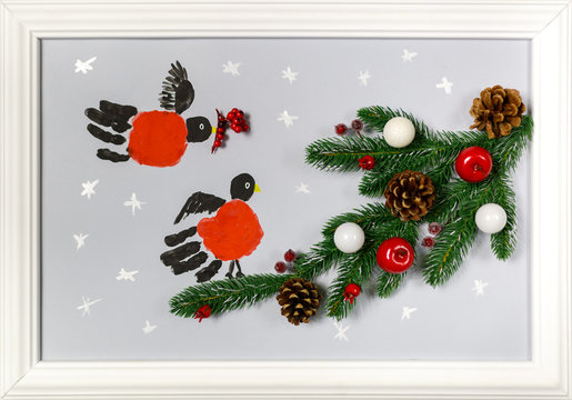 Prints of the hands palms are stylized as bullfinches. Branch of fir tree with cones, red apples and white balls. Handmade picture in a frame as an illustration of a New Year or Christmas holiday