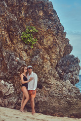 Attractive couple posing on a beach near big reef stones, enjoys a vacation on a beautiful island. 