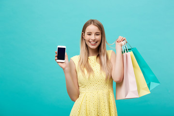 Portrait young attractive woman with shopping bags shows the phone's screen directly to the camera. Isolated on blue background