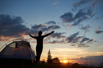 Rear view silhouette of female backpacker standing with open arms near a camping in the mountains at sunset under the evening yellow sky