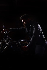 Night racer concept. Macho, brutal biker in leather jacket riding motorcycle at night time, copy space. Man with beard, biker in leather jacket sitting on motor bike in darkness, black background.