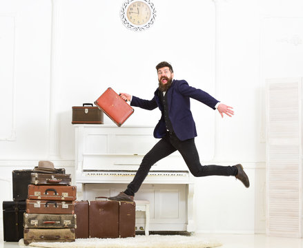 Macho attractive, elegant on cheerful face carries vintage suitcases, jumping. Man with beard and mustache in suit carries luggage, luxury white interior background. Long awaited vacation concept.