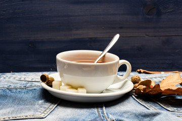 Mug filled with black brewed tea, spoon and autumn fallen leaves on denim background. Autumn drink concept. Tea served with spoon, sugar and decor acorn and leaves. Drink and acorn and oak leaves
