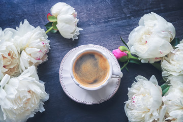 A cup of coffee and white peonies on dark wooden background. Top view, copy space. Toned image