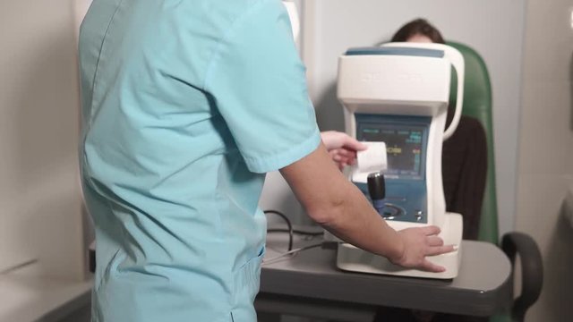 Optometrist is taking paper with medical examining result of vision from autorefractometer. Doctor is standing back to camera and patient is sitting behind apparatus