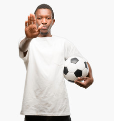 African black man holding soccer ball annoyed with bad attitude making stop sign with hand, saying...