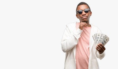 African man holding dollar bank notes thinking and looking up expressing doubt and wonder