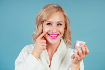 attractive and smiling blonde female person in a cotton white bathrobe applies moisturizer cream on face in the studio on a blue background . concept of skin care and spa