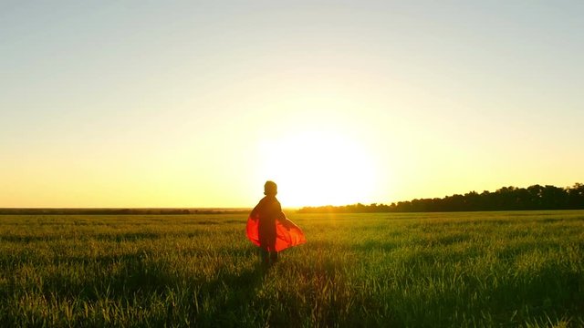 A child in a superhero costume runs on a green lawn against the backdrop of a sunset feigning flying, at a slow pace