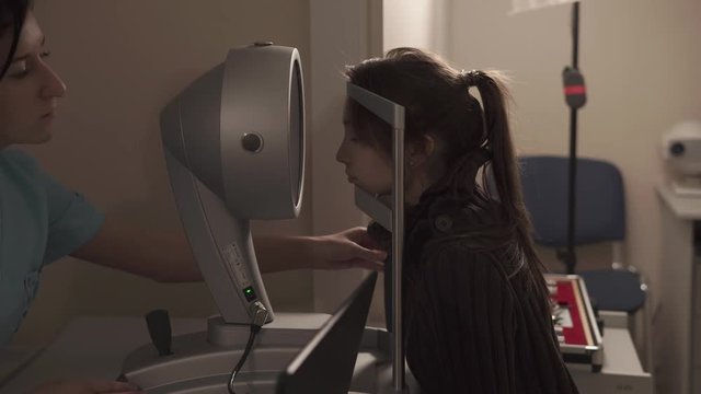 Side view shot of young girl placing her head on machine for eye topography. Female oculist helping her.
