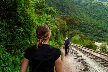 Poster Couple of young backpackers walking on the railway tracks towards Machu Picchu while being followed by a tourist woman © simonmayer
