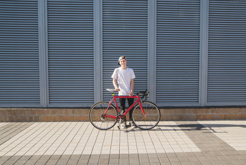 Student with a bicycle on the background of the wall. Happy young man in a white T-shirt and cap is standing on the background of a gray wall with a red bike and smiling