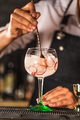 Three colorful cocktails in large wine glasses in a classic bar environment with dozens of blurry liquor bottles in the background.