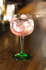 Three colorful cocktails in large wine glasses in a classic bar environment with dozens of blurry liquor bottles in the background.
