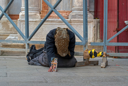 A homeless female beggar is begging on the street in Venice, Italy. A beggar woman holds a cardboard Cup in her hand for alms. The woman is wearing a brown scarf, a blue jacket and a long skirt