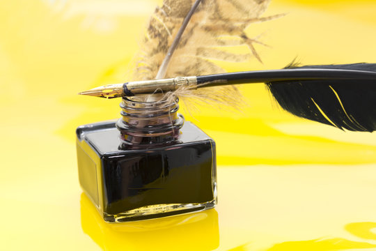 Close-up of quill on inkwell and feather on yellow background