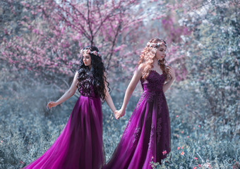 Obraz na płótnie Canvas Two sister girls are walking in the spring garden. The woman queen holds the hand of the girl princess. Same primkid, purple long dress, floral wreath. Sunny day, tree, green grass, flowering forest