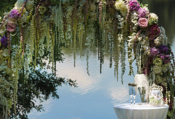 The wedding arch against the background of a water smooth surface decorated in flowers near her a little table with a ceremonial small pillow with wedding rings and glasses for champagne.