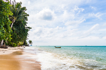 Tropical sandy beach with palms and a wooden boat in the background and blue ocean in the foreground during a bright sunny day, Haad Yao Beach, Koh Phangan, Thailand