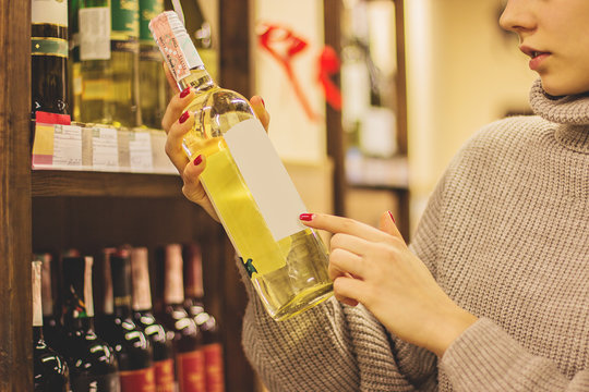 Close Up Of Young Woman Holding White Wine Bottle In Hands While Standing In Liquor Store. Girl Reading Or Pointing At Blank Label With Copy Space On Alcoholic Beverage Bottle At Winery Supermarket