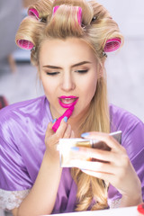 Beautiful female applying lip colour in front of pocket mirror. Charming young lady with curlers in her hair putting on very bright pink lipstick and holding mirror in her hand.