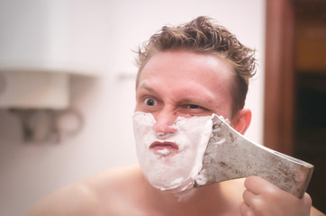 Frown man with brutal face is shaving with axe shaver in the bathroom.