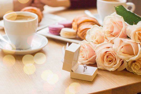 Close up of engagement ring in gift box, rose flower bouquet, coffee with croissant and macaroons on table at cozy cafe. Engagement of young couple in love. Marriage proposal concept. Ring for beloved