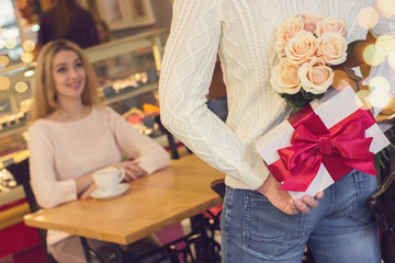 Back view man keeping flower bouquet and gift box behind his back for his beautiful girlfriend in cozy cafe. Man prepare unexpected surprise for his beloved woman. Valentines day, birthday concepts