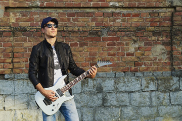 A young man in a leather jacket with a guitar against a brick wall