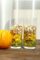 on a wooden table two glasses with pieces of fruit and muesli. next door is a fresh orange. vertical photo.