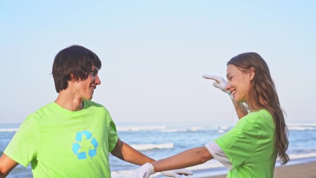 Happy young volunteers in green t-shirts with image recycle on an oceanic beach giving high five and smiling to camera after cleaning the beach. Volunteering and recycling concept.