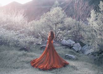 A brunette girl with a golden crown and in a red dress in a long train walking at sunset....