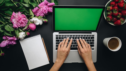 Female workspace. Woman tapping on the laptop computer keyboard with green screen. Bouquet of peony flowers, notebook and coffe on black background. Chroma key. Top view