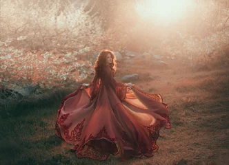 Ein brünettes Mädchen mit welligem, dickem Haar läuft zur Sonne und schaut zurück. Die Prinzessin hat ein luxuriöses, rotes Chiffonkleid, das im Wind flattert. Hintergrund eines feurigen Sonnenuntergangs und wilder Natur © kharchenkoirina