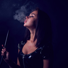 The portret of Young, beautiful girl smokes a hookah or shisha or kalian. Smoke and passion. Leather or latex dress, sexy outfit.The pleasure of smoking.