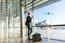 Young casual female traveler at airport, holding smart phone device, looking through the airport gate windows at planes on airport runway.
