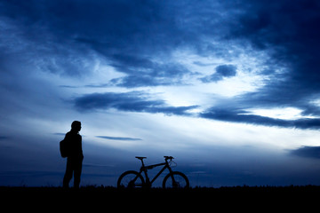 Obraz premium Man’s silhouette stands near his mountain bike and looks forward against dark blue sky
