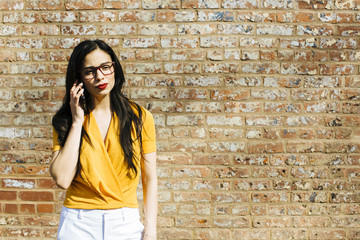 Portrait of a young brunette woman talking on the phone in in front of brick wall, with text space on side