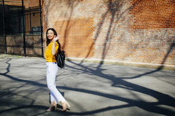 Portrait of a young woman with backpack walking in front of a brick building, waving at camera