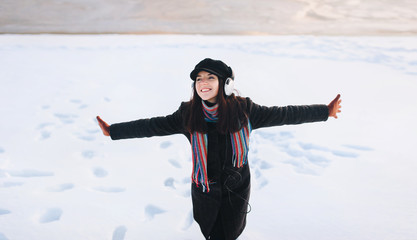 Fototapeta na wymiar Young funny girl in headphones in winter on a snow background. Playlist with your favorite songs. To cheer up the music. Spreading hands. Copy space.