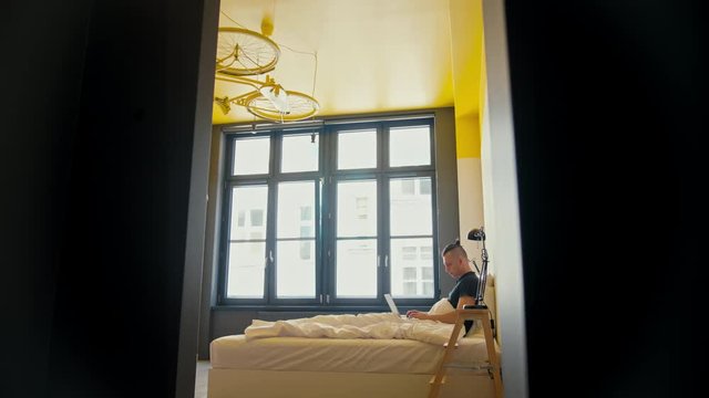 Young Man is Lying in a Bed with Laptop at Home in the Morning in a Cozy Apartment Room with Big Window