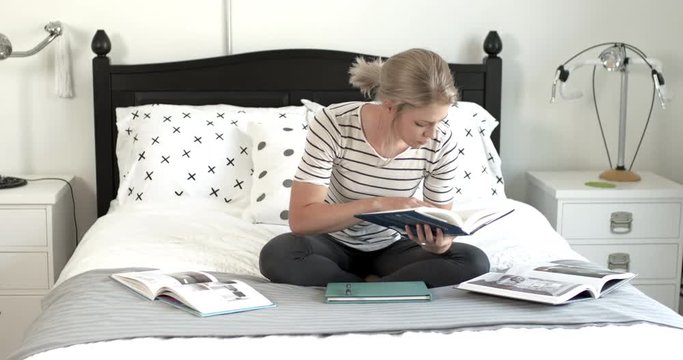 Young woman studying, reading, writing, drawing, relaxing on bed in apartment or dorm