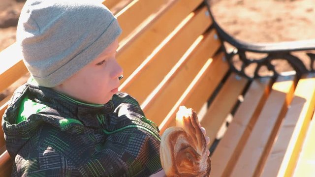 Boy is sitting on a bench in the park in the spring and eating a bun with sugar. Side view.