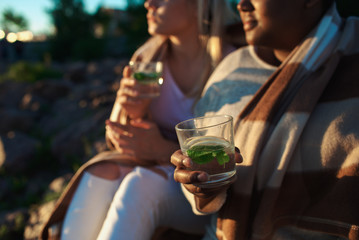 Two crop diverse women holding glasses of fresh drinks while sitting under warm blanket during multiethnic party. 