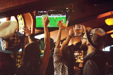 group or company of friends - young guys and girls holding glasses of beer, watching football,...