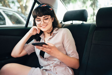 Beautiful young pretty girl is using a smart phone and smiling while sitting on back seat in the car