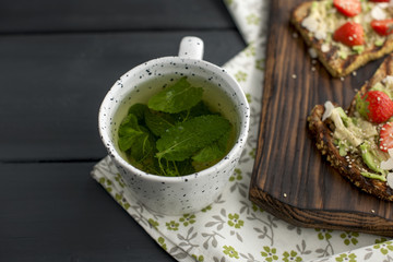 tea with fresh mint in a white mug on a napkin and black wooden background, healthy breakfast with toast. Copy space.