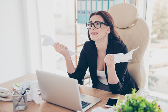 Portrait of crying exhausted woman in glasses holding crumpled lists in hands, business in not successful, hard days, tired from routine, having computer laptop on the table, looking away