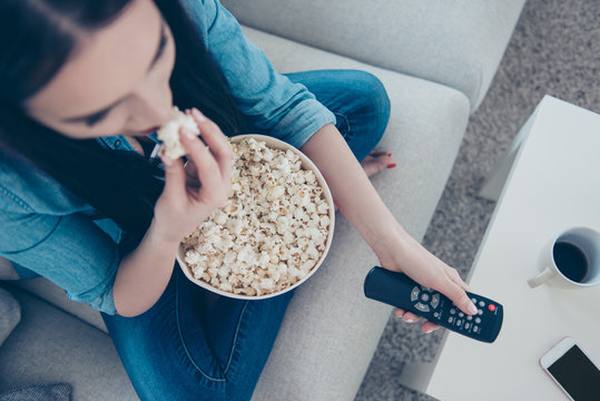Top view portrait of woman in jeans outfit sitting with crossed legs on sofa searching movie using remote controller having bucket with pop corn sitting in living room