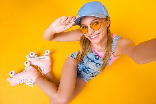 Top view self portrait of positive slim girl with beaming smile shooting selfie on front camera while sitting on ground floor in roller skates isolated on yellow background